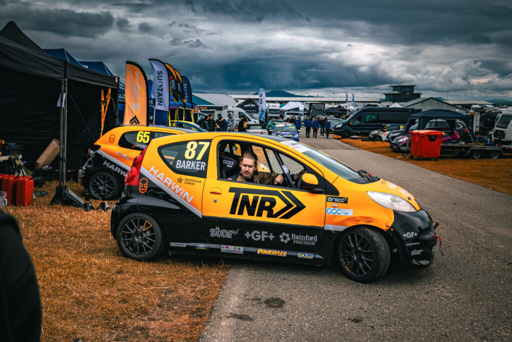 A man is driving a motorsport vehicle near a pit stop