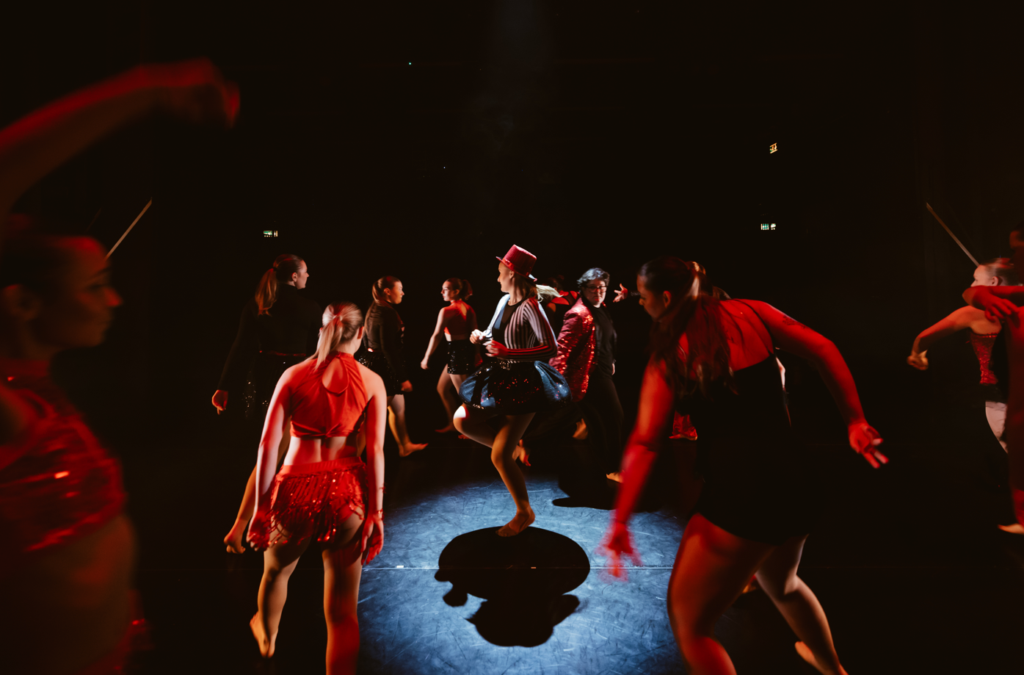 A large group of students, dressed in costume, dancing on stage together under a spotlight